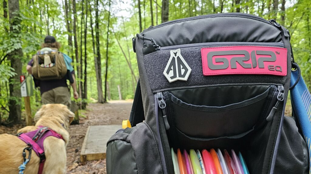 A gray GRIPeq CX1 bag sitting on a tee pad with a person walking away from it carrying a sand BX3 bag and a dog in between them.