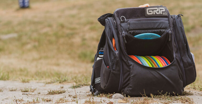 A black AX5 disc golf bag sits on the ground at a course. The front flap is open and tucked into the bag revealing the discs inside. There is a water bottle in the pocket and a towel is showing in the side pocket.
