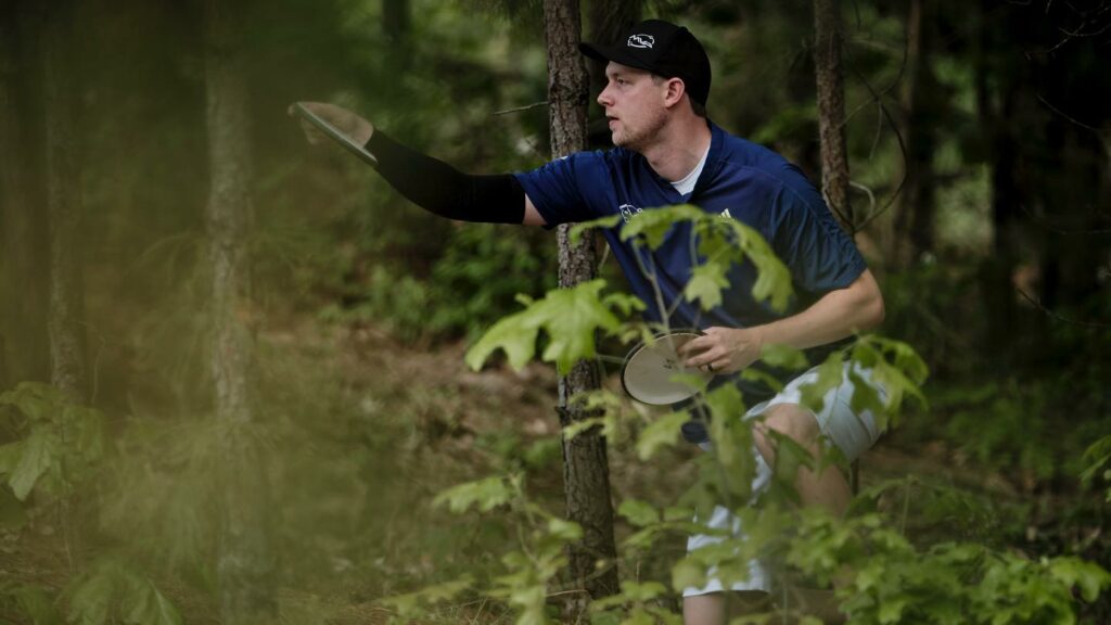 Team GRIPeq member Simon Lizotte kneels in the forest with a disc in hand, preparing to putt.