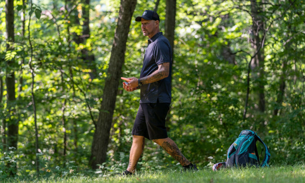 Patrick Brown walks across the course with a disc in-hand. His turquoise GRIPeq AX5 bag sits on the ground behind him.
