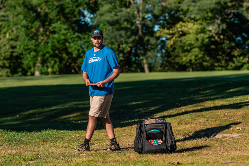 Brian Earhart prepares to throw a disc with a black GRIPeq disc golf bag at his feet. The front flap is open showing his discs inside.