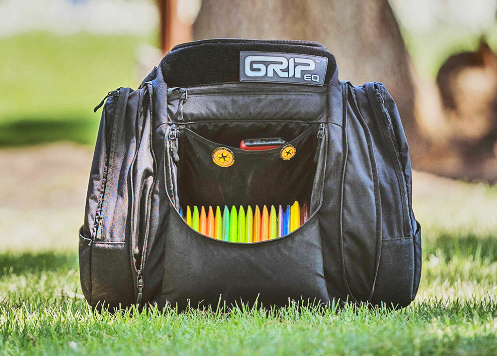 A black GRIPeq AX5 bag sits in the grass. The front flap is open and the discs inside are visible.