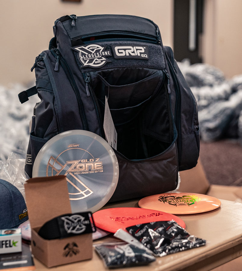 A black GRIpeq BX3 bag sits on a table indoors, surrounded by 3 discs, a belt, and a hat.