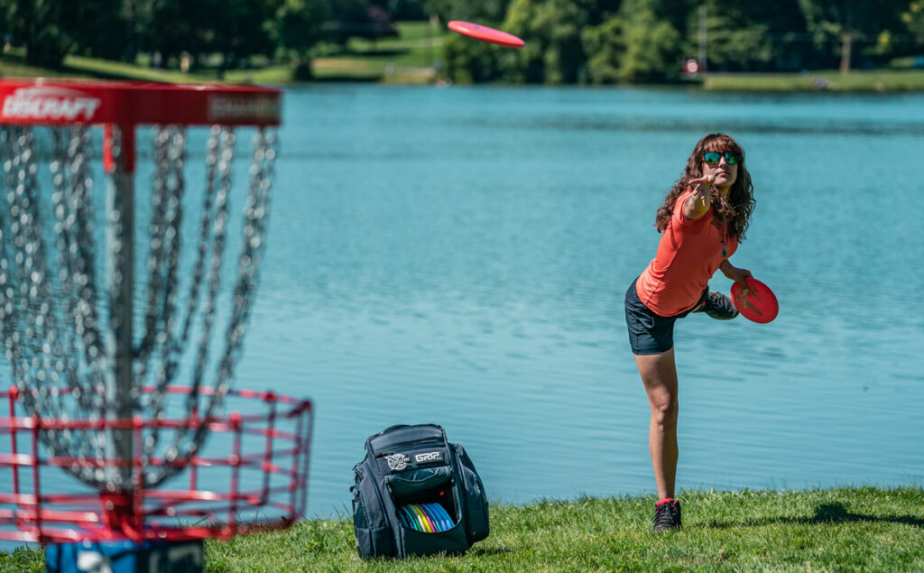 Team GRIPeq member Jessica Weese putting into a disc golf basket with the disc in the air, mid-throw. There is pond behind her and her GRIPeq disc golf basket sits on the ground next to her.