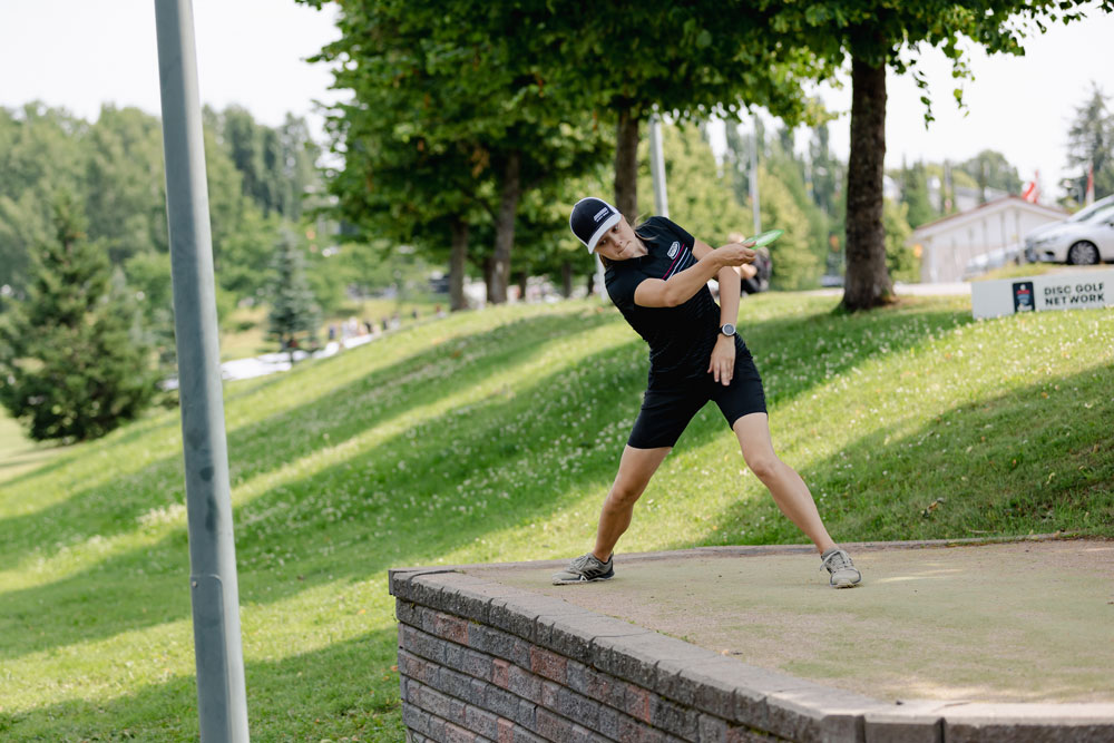Henna Blomroos stands on the tee and reaches back to throw a disc golf drive.