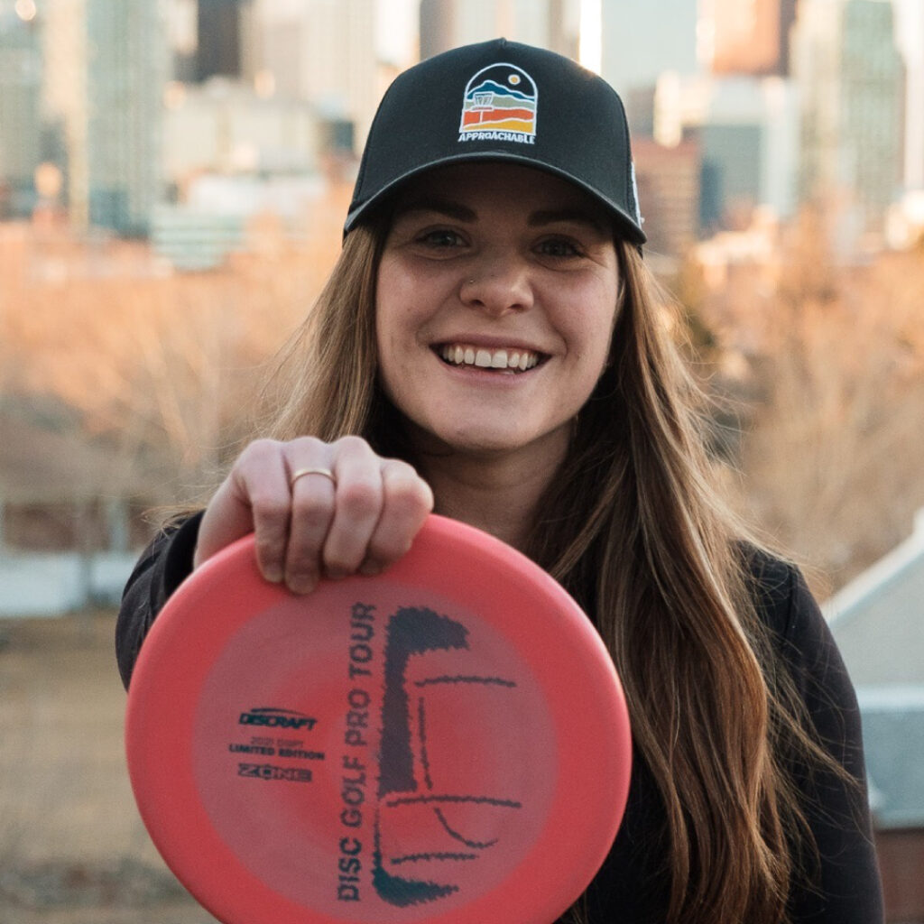 Jo Henderson holding a disc at the camera and smiling. She's wearing an Approachable Podcast hat.