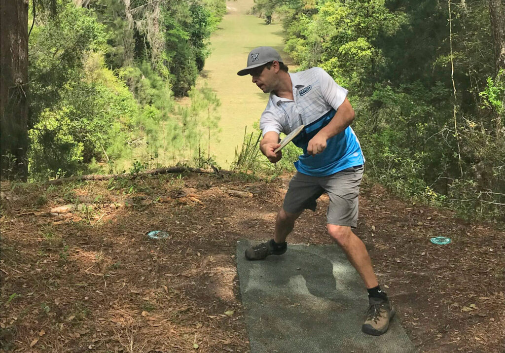 Steve Brinster winds back to throw a backhand disc golf tee off up on top of a small cliff.