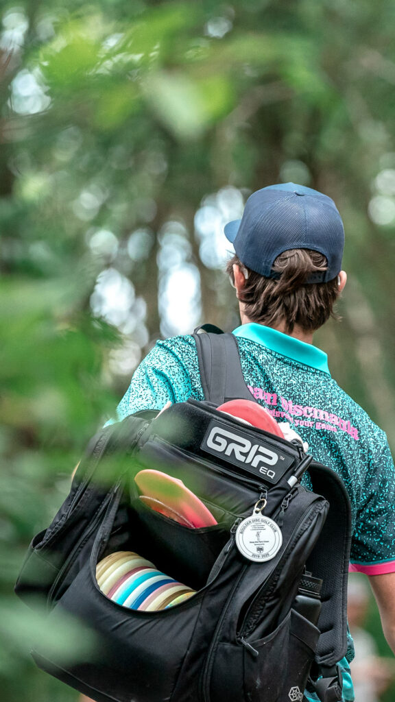 A back view of someone wearing a black GRIPeq AX5 bag on their back. There is a keychain hanging from one of the zippers and the bag is full of discs with the front flap open and tucked inside.
