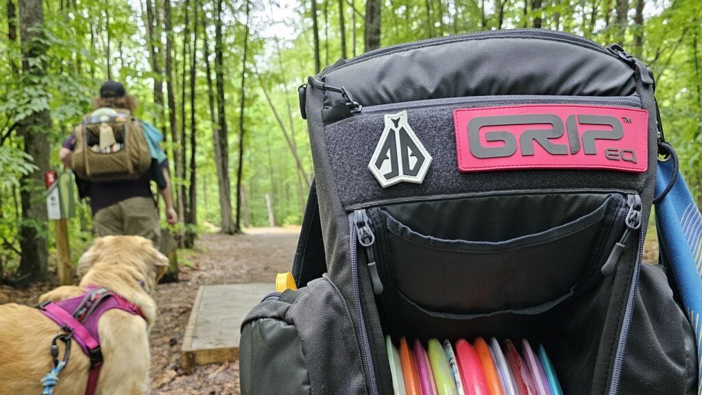 Gray CS2 bag with a Pink GRIPeq patch and an Anthony Barela patch. You can see a disc golf fairway, disc golfer, and dog in the background.