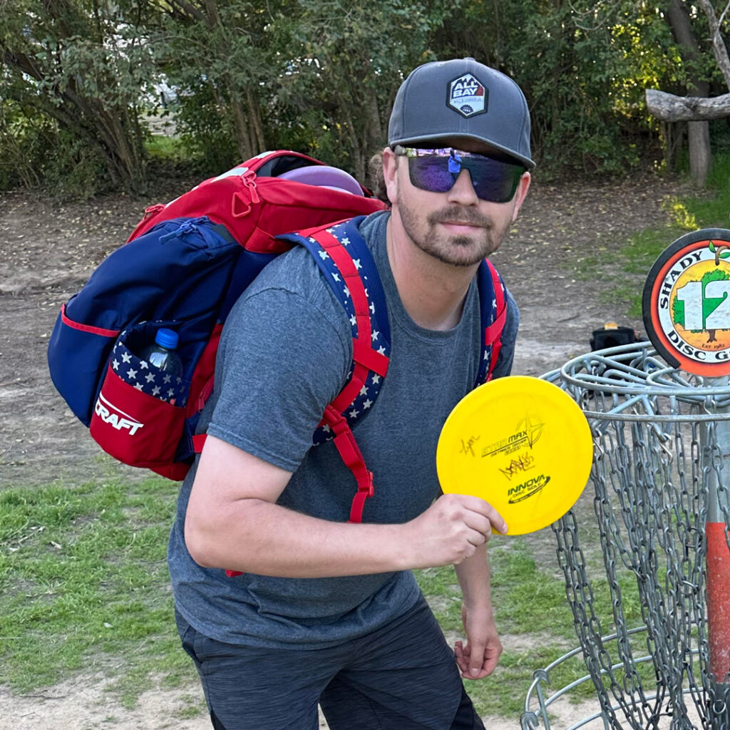 Team GRIPeq member Sam Minges stands next to a disc golf basket, holding a disc.