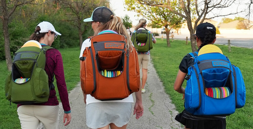 3 GRIPeq pros carry GRIPeq disc golf bags on their backs. The image shows them from the back walking down a path.