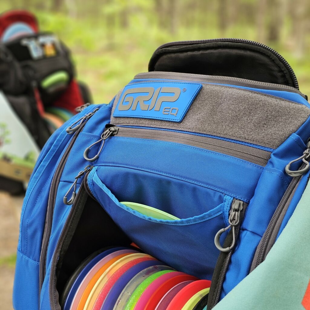 AX6 Ocean blue bag close up with MB-PX1 bag in the background. Bags are sitting on a bench at a disc golf course.